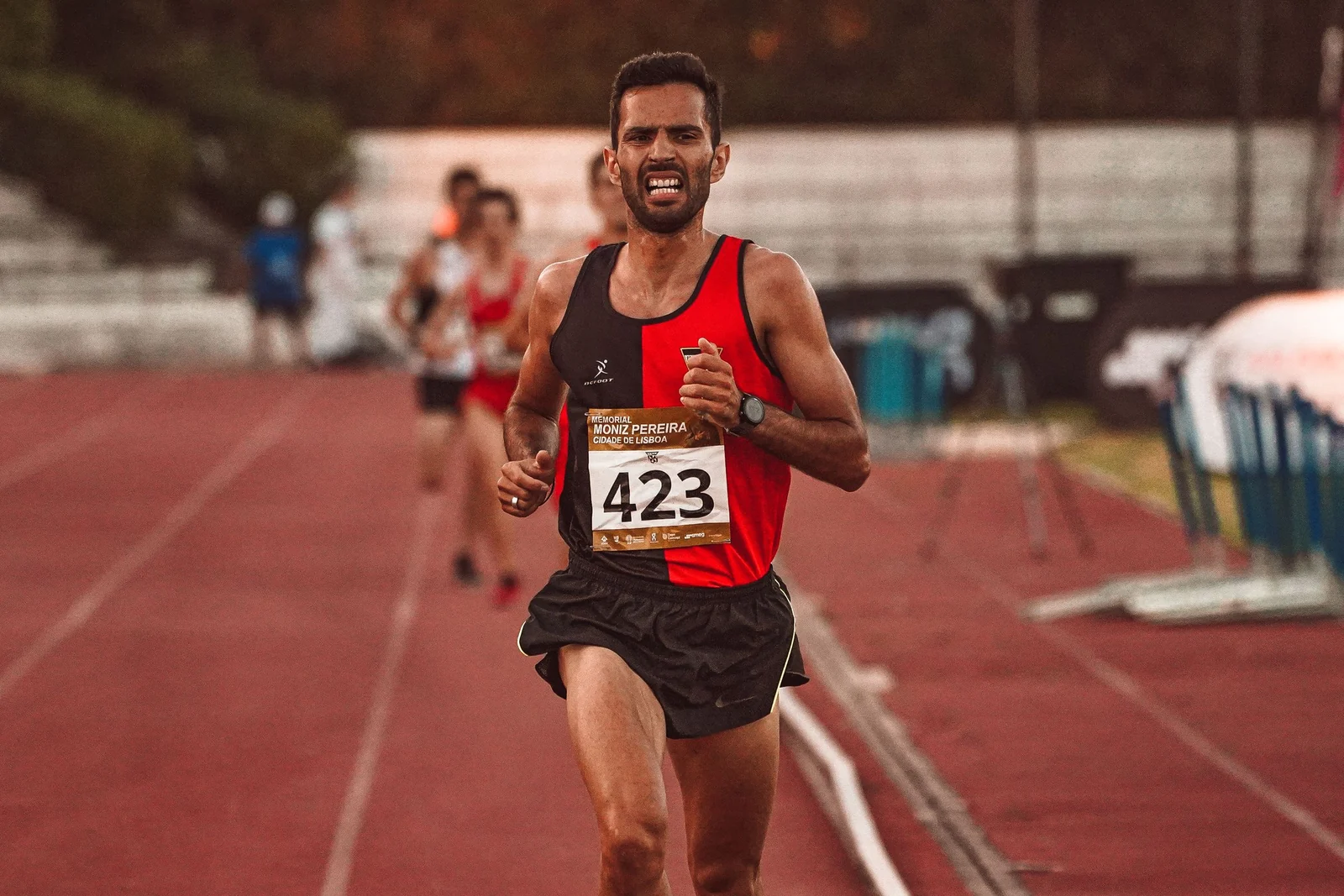 A runner in a red and black racing vest and black shorts competing on an outdoor track.