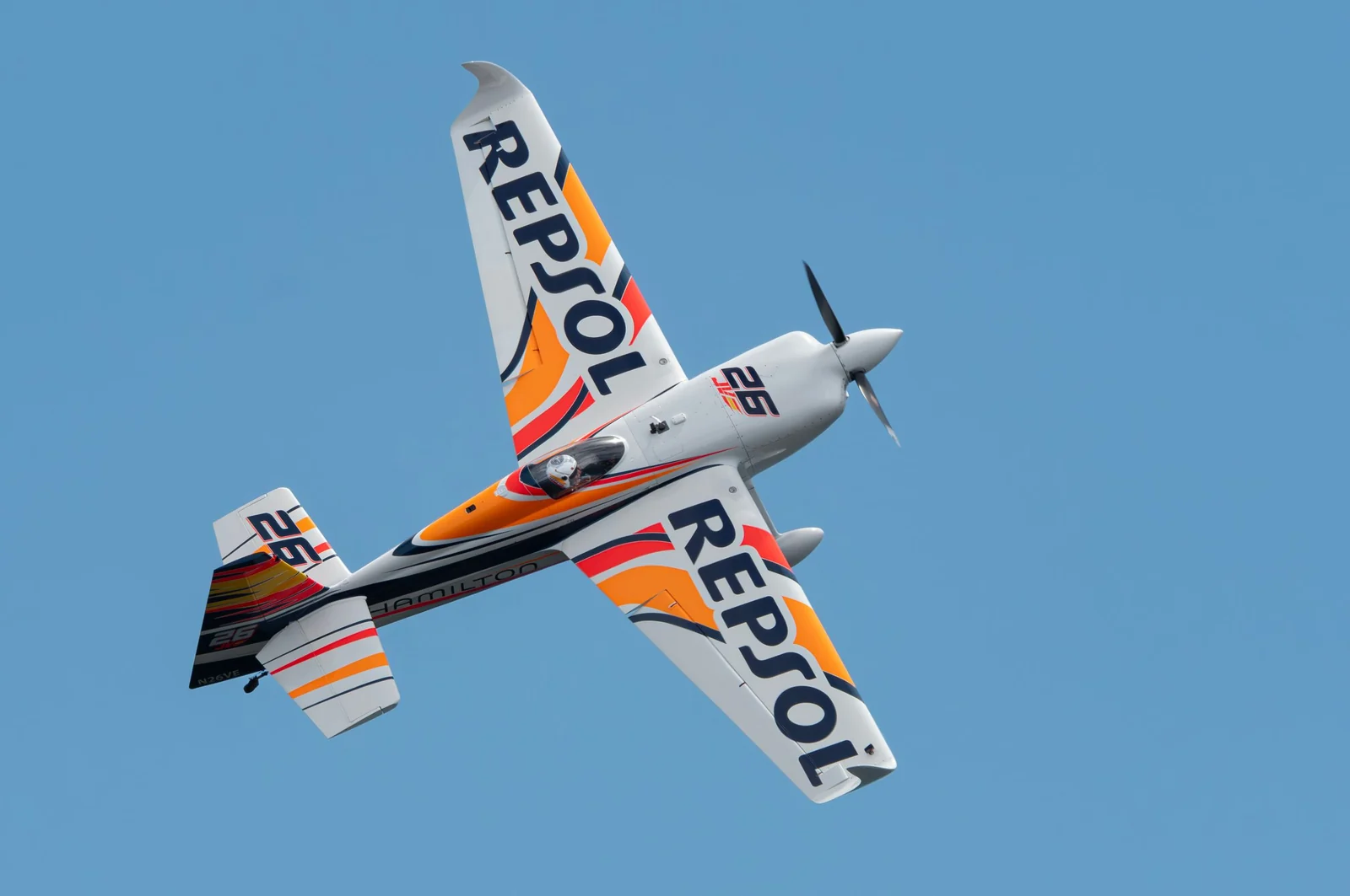 An aerobatic propeller plane banking steeply in flight against a clear blue sky.
